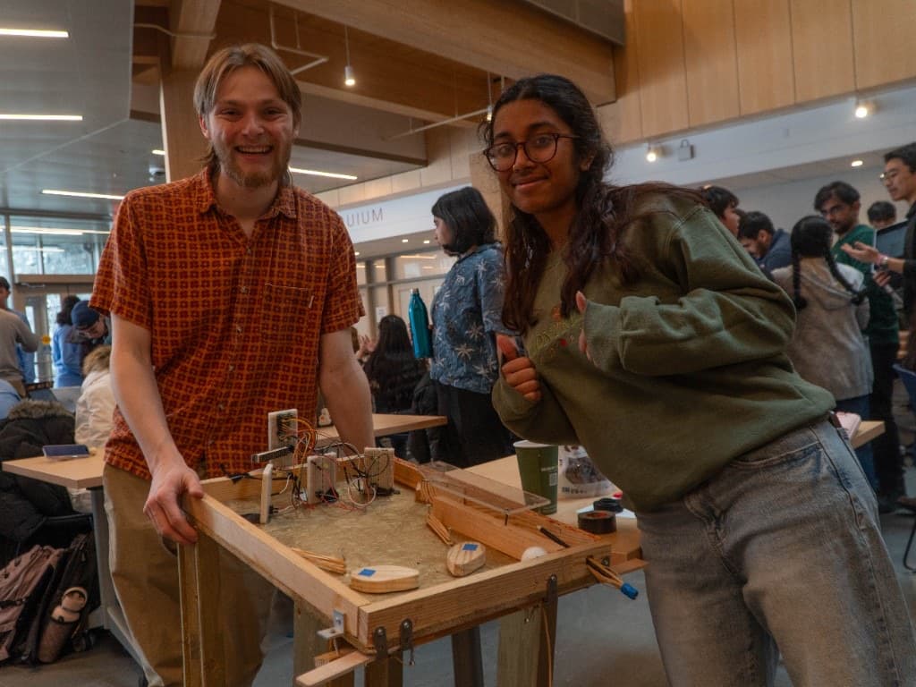 Two builders smiling with a handmade wooden hardware game in the atrium.