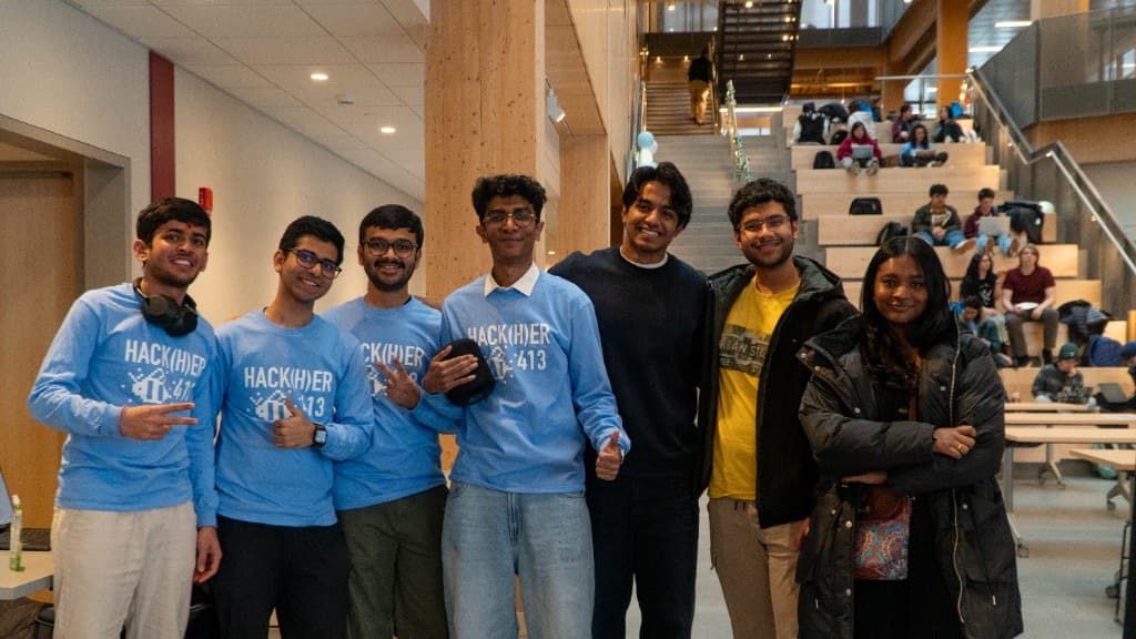 A group of attendees in Hack(H)er413 shirts posing in front of the wooden grandstand seating.
